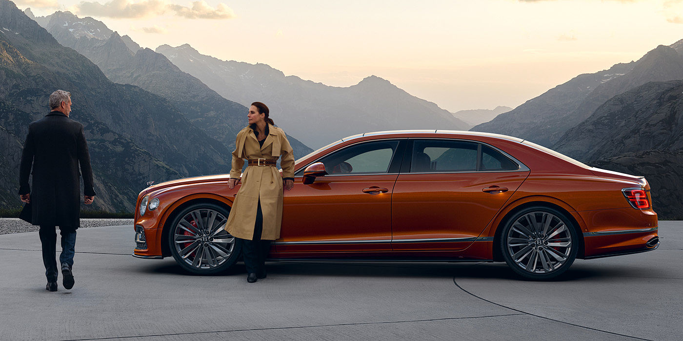 Bentley Hatfield Bentley Flying Spur Speed parked in Orange Flame coloured exterior parked, with mountainous background and two people in view.