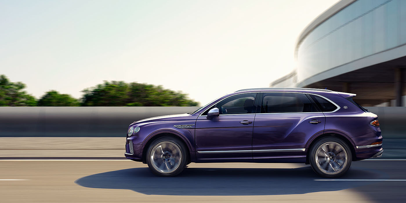Bentley Hatfield Bentley Bentayga Extended Wheelbase Mulliner luxury SUV profile in Tanzanite Purple paint, driving on an country road past trees.