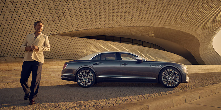 Bentley Hatfield Bentley Flying Spur Mulliner sedan in Rose Gold over Magnetic duotone paint parked by a curved wall with person in suit jacket standing on the left